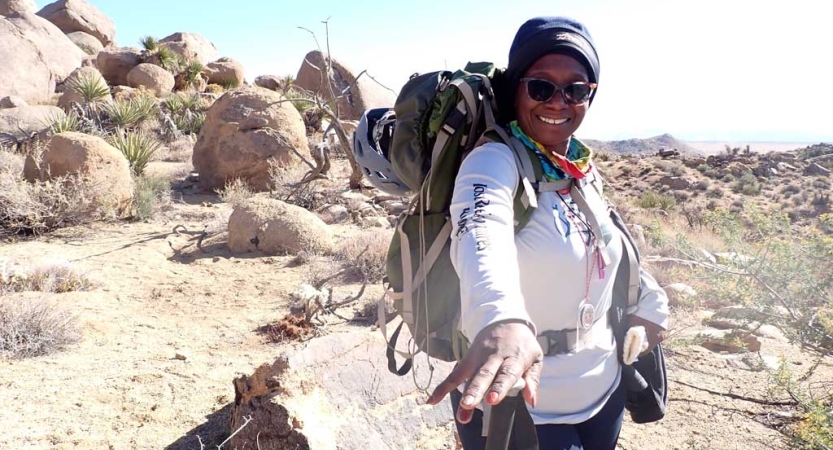 A person wearing a backpack smiles and stretches a hand toward the camera. They are standing in a desert landscape. 